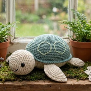 Pode incluir: Uma tartaruga de crochê com uma carapaça azul clara e uma cabeça e membros cor de creme. A tartaruga está sentada em uma cama de musgo verde. A tartaruga tem um rosto sorridente.