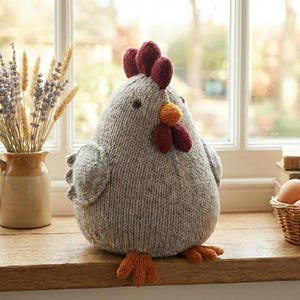Peut inclure: Un poulet tricoté gris avec une crête rouge, un bec orange et des pattes orange est posé sur une surface en bois. Le poulet fait main est à côté d'un vase de fleurs séchées et d'un panier d'œufs. La fenêtre en arrière-plan fournit la lumière naturelle.