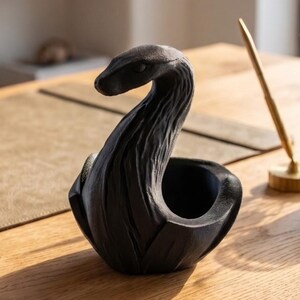 Könnte beinhalten: Eine mattschwarze, skulpturale Schale in Form einer zusammengerollten Schlange mit einer texturierten, holzähnlichen Oberfläche. Der erhobene Schlangenkopf bildet den Hals des Gefäßes, während der Körper eine hohle, offene Schale für kleine Gegenstände formt.