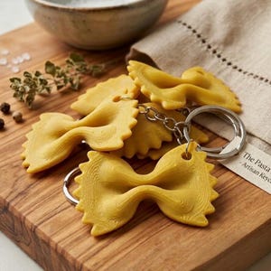 Pode incluir: Close-up de vários chaveiros amarelos em forma de massa farfalle com argolas prateadas. Os chaveiros estão numa tábua de cortar de madeira com uma tigela pequena e um guardanapo de linho. O texto no guardanapo diz "The Pasta - Artisan Keych".