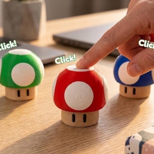 Può includere: Tre giocattoli da scrivania a forma di fungo, rossi, verdi e blu, ciascuno con dettagli circolari bianchi. Il testo "Click!" è visibile. La base di ogni fungo è marrone chiaro. Un dito preme uno dei funghi.