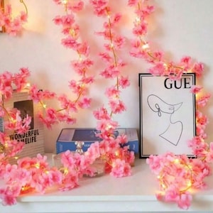 May include: Pink cherry blossom string lights draped over a shelf with books and a framed print. The print has the word "GUE" and a line drawing of a woman's face. The lights are wrapped around the blossoms.