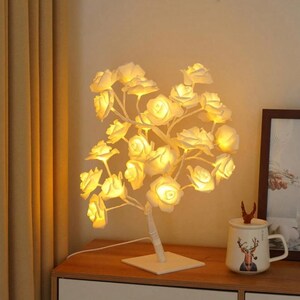 Pode incluir: Uma lâmpada de árvore de rosas brancas com luzes LED amarelas quentes. A lâmpada tem uma base quadrada e um design de tronco de árvore com inúmeras luzes em forma de rosa. A lâmpada está sobre uma superfície de madeira.