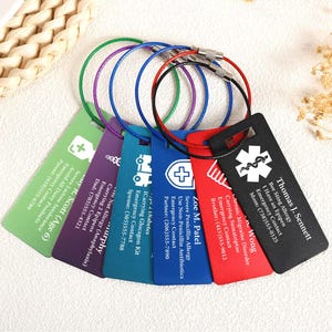 May include: A selection of brightly coloured medical alert tags, each displaying different medical conditions and emergency contact details. The tags are rectangular with rounded corners and attached to a metal loop. The tags are in green, purple, blue, red, and black.
