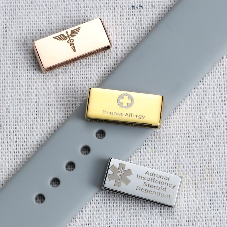 May include: Three rectangular metal medical alert charms for a watch band. One rose gold charm features a caduceus symbol. One gold charm displays a cross and the text Peanut Allergy. One silver charm shows a Star of Life and Adrenal Insufficiency Steroid Dependent.