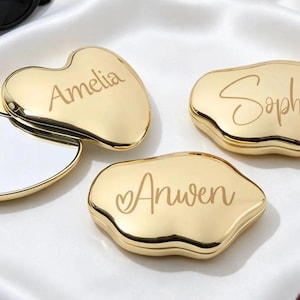 May include: Three gold-colored, irregular-shaped compact mirrors with smooth, reflective surfaces. One mirror is open, revealing the glass, while two are closed. The names Amelia, Sophia, and Anwen are engraved in a cursive script on the lids.