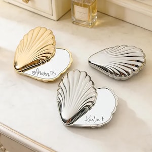 May include: Three shell-shaped compact mirrors with metallic finishes. One gold mirror is open, displaying the name Anwen with two hearts. One silver mirror is open, showing the name Kaelin with a lightning bolt. One silver mirror is closed.