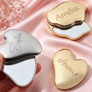 May include: Three heart-shaped compact mirrors with metallic finishes. One silver mirror is held, engraved with the name Sophia. Two gold mirrors rest nearby, engraved with the names Amelia and Anwen. Each mirror features a smooth, reflective surface.