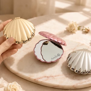 Pode incluir: Três espelhos compactos em forma de concha com acabamentos metálicos dourado, prateado e rosa. O espelho rosa está aberto, revelando uma superfície refletora com o texto Amelia e um símbolo de estrela. Cada estojo tem um design exterior canelado.