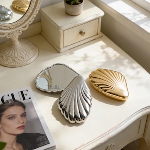May include: A silver and gold seashell-shaped compact mirror with a scalloped edge. The silver mirror has the name "Thalia" engraved on the mirror. A Vogue magazine is in the foreground. The mirrors are on a white dressing table.