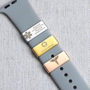 May include: A grey silicone watch strap featuring three rectangular metal medical alert charms. The top silver charm displays a Star of Life symbol and the text Adrenal Insufficiency Steroid Dependent. The middle gold charm shows a cross and Peanut Allergy text. The bottom rose gold charm features a caduceus symbol.