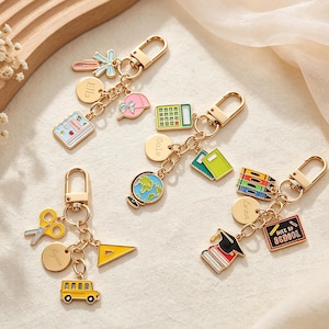 May include: Four gold-toned metal keychains featuring school-themed enamel charms. Each keychain includes a personalized circular disc, a lobster clasp, and various charms such as a globe, calculator, school bus, scissors, crayons, and a chalkboard.