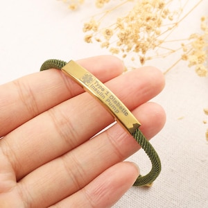 May include: Olive green medical alert bracelet with a gold-coloured rectangular plate. The plate is engraved with the words "Type 1 Diabetic Insulin Pump". The bracelet is held in a hand.