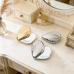 May include: Three seashell-shaped compact mirrors in gold and silver tones, with personalized names engraved on the mirror surface. The mirrors are open, revealing the reflective surface. The background includes a vanity and decorative items.