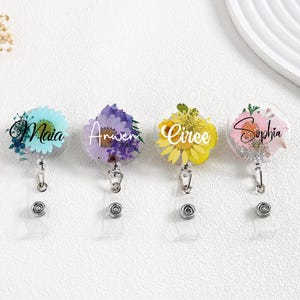 May include: Four clear, round badge reels with floral designs and personalized names: Maia, Anwen, Circe, and Sophia. Each reel features a different colour scheme, including blue, purple, yellow, and pink flowers, with a retractable cord and a metal clip.