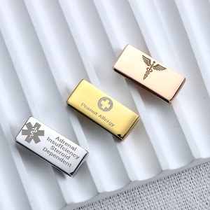 May include: Three rectangular metal medical alert tags in silver, gold, and rose gold tones. The silver tag features a Star of Life symbol and the text Adrenal Insufficiency Steroid Dependent. The gold tag shows a cross and Peanut Allergy text.