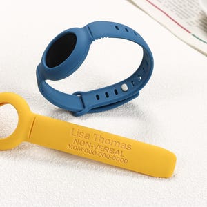 May include: A blue silicone wristband with a black oval centre and a yellow silicone band with the text "Lisa Thomas NON-VERBAL MOM:000-000-0000". The wristband is designed to be worn on the wrist.