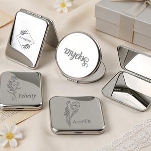 Può includere: Una collezione di specchietti da borsa in metallo argentato di forma quadrata e rotonda. Alcuni specchietti sono chiusi e mostrano disegni floreali incisi e nomi come Amelia, Felicity e Sophia, mentre altri sono aperti.