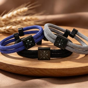 Op de afbeelding: Drie armbanden met dubbel koord in blauw, grijs en zwart. Elk heeft een vierkant zwart metalen middendeel gegraveerd met een kruisontwerp, letters en kleine iconen zoals een klavertje, palmboom of pootafdruk. De koorden zijn geweven.