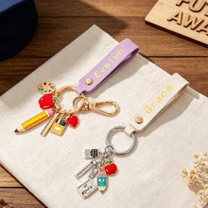 Peut inclure: Deux porte-clés avec des lanières personnalisées. L'un est lilas avec "Evelyn" en lettres dorées, l'autre est crème avec "Grace". Les breloques comprennent un crayon, une pomme, une palette de peinture et des fournitures scolaires. Le fond est une surface en bois.
