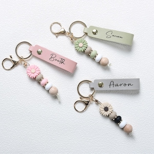 Op de afbeelding: Vier gepersonaliseerde sleutelhangers met goudkleurige hardware. Elke sleutelhanger heeft een leren label met een naam: "Booth", "Saron" en "Aaron". De sleutelhangers hebben decoratieve kralen en bloemaccenten in verschillende kleuren, waaronder roze, groen en neutrale tinten.