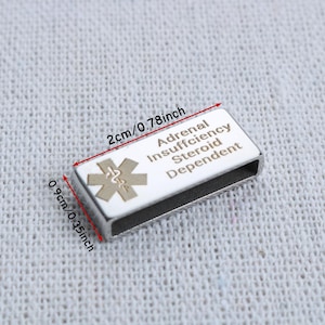 May include: A rectangular silver-tone medical alert tag with a white enamel inlay featuring a gold-colored Star of Life symbol and the text Adrenal Insufficiency Steroid Dependent. The tag measures 0.78 inches by 0.35 inches as indicated by red arrows.