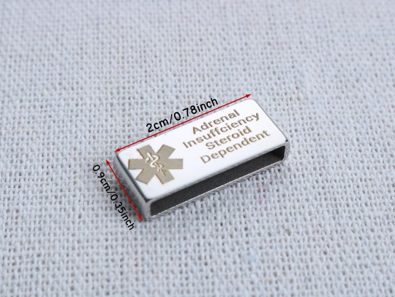 May include: A rectangular silver-tone medical alert tag with a white enamel inlay featuring a gold-colored Star of Life symbol and the text Adrenal Insufficiency Steroid Dependent. The tag measures 0.78 inches by 0.35 inches as indicated by red arrows.
