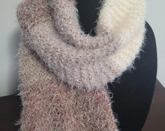May include: A soft, fluffy scarf in shades of cream, light brown, and mauve. The scarf is draped over a black mannequin head, showcasing its texture and color variations. The knitted design appears warm and cozy.