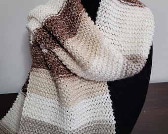 May include: A hand-knitted scarf displayed on a black mannequin head. The scarf features horizontal stripes in shades of white, brown, and beige. The knitted texture is clearly visible, showcasing the craftsmanship of the piece. The scarf is designed to be worn around the neck.