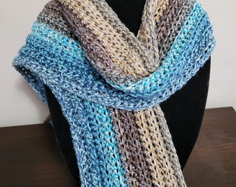 May include: A handmade crochet scarf in shades of blue, gray, and tan. The scarf is draped over a black mannequin head, showcasing its textured stitch pattern and color gradient. The scarf appears to be made of soft yarn.