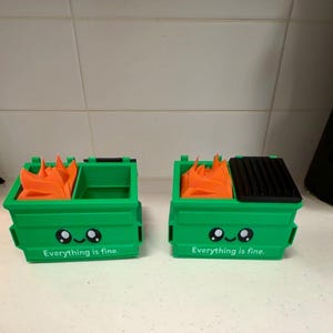 Op de afbeelding: Twee groene miniatuur vuilnisbakfiguren met oranje vlammen erin. Elke vuilnisbak heeft een lachend gezicht en de tekst "Everything is fine". Eén vuilnisbak heeft een zwart deksel.