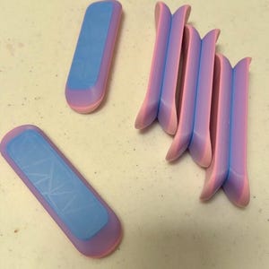 May include: A collection of small, brightly coloured 3D-printed items are displayed on a light surface. The objects are a mix of blue and pink, with a rectangular form and a textured finish. Some are curved, while others are flat.