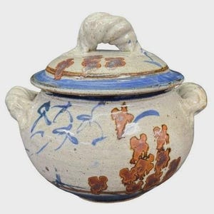 May include: A ceramic lidded jar with a speckled off-white base, adorned with blue and rust-colored floral designs. The lid features a blue rim and a textured handle. The jar has two side handles.