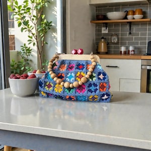 May include: A blue crochet handbag with a wooden frame and handle. The bag features colorful floral squares in shades of pink, yellow, and blue. The handle is made of large wooden beads. Two red decorative pieces are on top of the wooden frame.