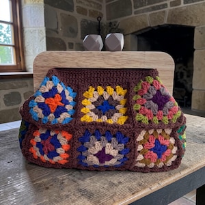 May include: A handmade crochet clutch with a wooden frame. The bag features a brown base with colorful granny squares in shades of blue, orange, yellow, green, and purple. The wooden frame has two gray geometric accents.