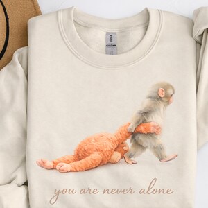 May include: Cream-colored sweatshirt featuring a watercolor illustration of two monkeys. One monkey is walking, holding the other, orange-colored monkey. The text "you are never alone" is printed in a script font.