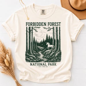 May include: A cream-colored short-sleeve t-shirt featuring a dark green graphic print of a forest scene with a unicorn, a moon, and the text "FORBIDDEN FOREST NATIONAL PARK EST. 990 AD" centered on the front of the garment.