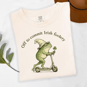 May include: Cream-colored t-shirt with the phrase "Off to commit Irish foolery" printed in dark green. The shirt features a cartoon frog wearing a hat and holding a shamrock while riding a scooter. The shirt has a Comfort Colors tag.