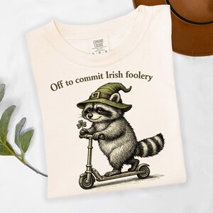 May include: Cream-colored t-shirt with the text "Off to commit Irish foolery." The graphic features a raccoon wearing a green hat and holding a shamrock, riding a scooter. The shirt is folded.
