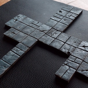 Op de afbeelding: Een donkergrijs, kruisvormig object gemaakt van individuele, verweerde rechthoekige blokken. De blokken hebben een ruwe, getextureerde uitstraling met zichtbare scheuren en onvolkomenheden. Het object ligt op een zwart, getextureerd oppervlak.