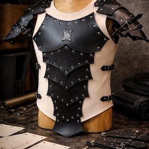 Puede incluir: Armadura de cuero negro y crema exhibida en un maniquí de madera. La armadura presenta detalles remachados, hombreras y correas ajustables. Un pequeño logotipo es visible en el pecho. La armadura está diseñada para protección y cosplay.