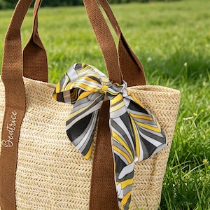 May include: A woven straw tote bag with brown straps and trim. A patterned scarf in yellow, gray, and black is tied to one of the straps. The word "Beatrice" is written on the side of the bag.