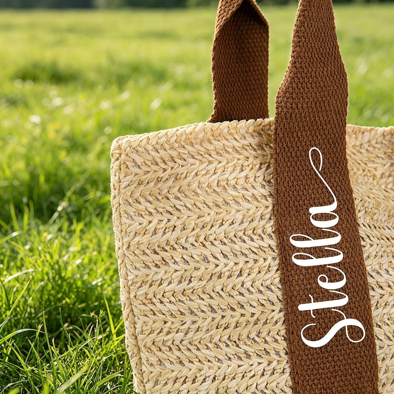 May include: A woven straw tote bag with brown straps. The word "Stella" is written in white script on one of the straps. The bag is set against a background of green grass.