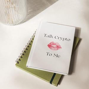 Peut inclure: Deux cahiers, un à spirales et un à couverture rigide, sont empilés sur une surface blanche. Le cahier supérieur porte le texte "Talk Crypto To Me" avec une image de lèvres roses. Le cahier inférieur est vert olive.