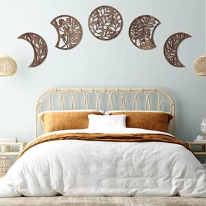 Peut inclure: Décoration murale en bois représentant les phases de la lune avec des motifs de feuilles et de branches. Les pièces marron sont montées sur un mur bleu clair au-dessus d'un lit avec une literie blanche et des oreillers couleur moutarde. Décoration de chambre.