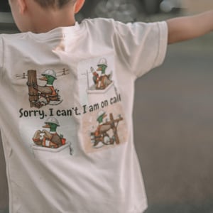 May include: White t-shirt with a cartoon duck design. The design features ducks in construction worker attire, with the text "Sorry, I can't. I am on call." The shirt is short-sleeved.
