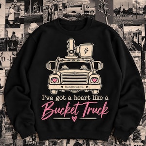 May include: A black crewneck sweatshirt featuring a graphic of a bucket truck with pink heart-shaped headlights and a lightning bolt icon on the bucket. The text reads "I've got a heart like a Bucket Truck" and "BuckIDTruck Co." in white and pink.