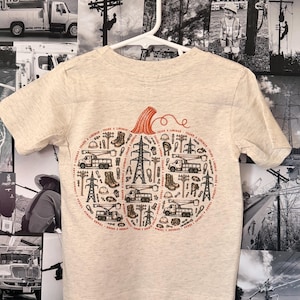 May include: A heathered beige short-sleeve t-shirt featuring a large pumpkin-shaped graphic composed of black and red illustrations of utility trucks, power poles, boots, and hard hats, with the text THANK A LINEMAN repeated along the border.