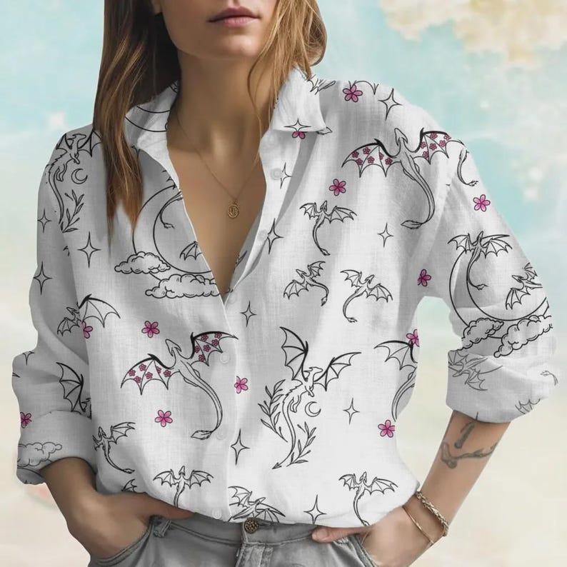 May include: White button-down shirt with a repeating pattern of black dragons, moons, stars, and pink flowers. The long-sleeved shirt has a classic collar and is made of a lightweight fabric.