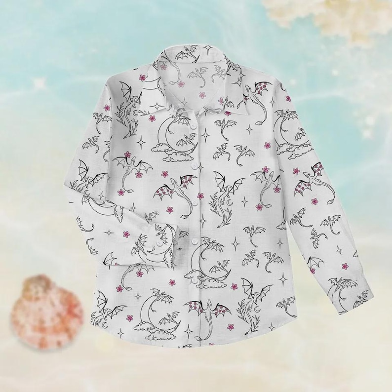 May include: White button-down shirt with a repeating pattern of black dragons, moons, stars, and pink flowers. The long-sleeved shirt has a classic collar and buttoned cuffs. The shirt is made of a lightweight fabric.